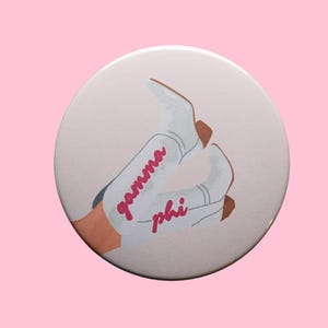 May include: A round button with a light pink background featuring an illustration of white cowboy boots. The boots have the words "gamma phi" in pink script. The button is set against a pink background.