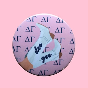 May include: A round button with a pink background featuring a pattern of Greek letters and a pair of white cowboy boots. The boots have the words "dee gee" in a cursive font.