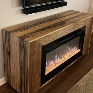 May include: A wooden entertainment center with a built-in electric fireplace. The fireplace has a black frame and a realistic flame effect. The entertainment center is made of dark wood with a natural grain pattern. A flat screen TV is mounted above the entertainment center.