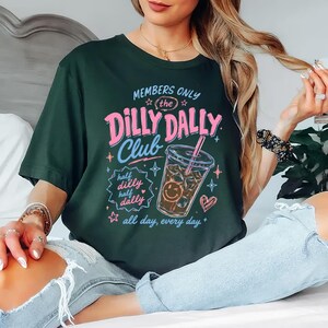 May include: A dark green t-shirt featuring a graphic print with the text "MEMBERS ONLY the DILLY DALLY Club half dilly half dally all day, every day." The design includes a pink and blue iced coffee cup with a smiley face, stars, and a heart.