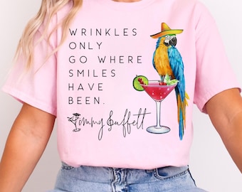 May include: Pink t-shirt featuring a parrot wearing a sombrero and holding a cocktail. The text on the shirt reads "Wrinkles only go where smiles have been. Jimmy Buffett"