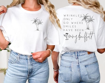 May include: White t-shirt with a palm tree graphic on the front and the text "Wrinkles Only Go Where Smiles Have Been" with a signature and cocktail graphic on the back. The shirt is paired with blue jeans.