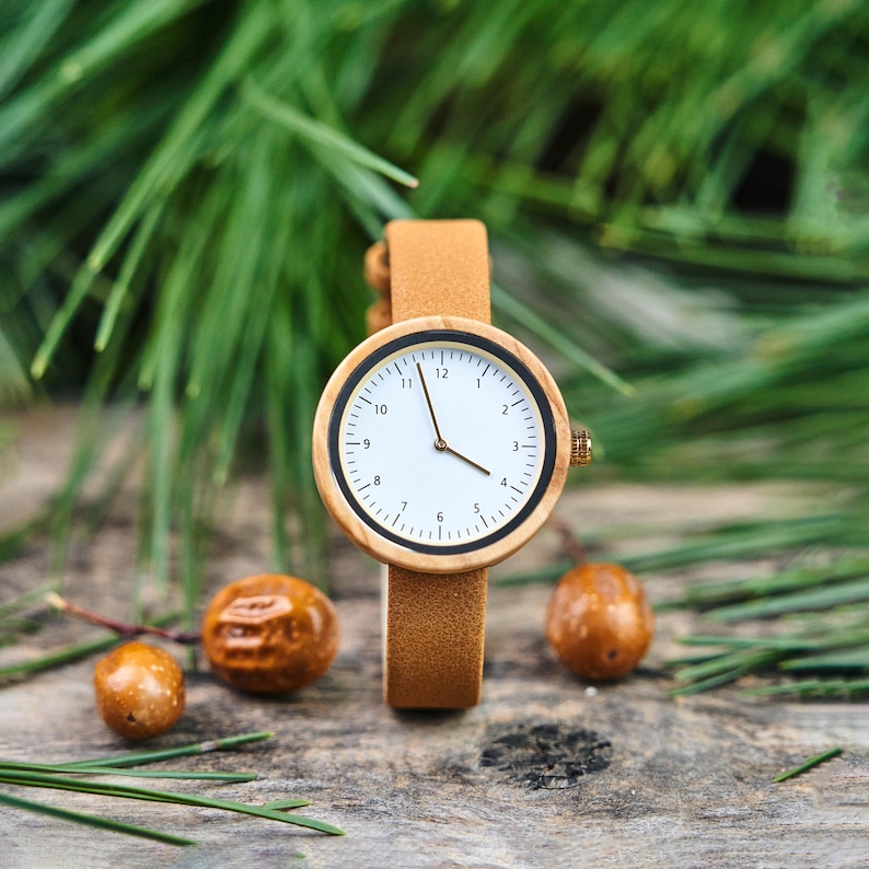K&ouml;nnte beinhalten: Eine Armbanduhr mit Holzgeh&auml;use und hellbraunem Lederarmband. Das Zifferblatt ist wei&szlig; mit schwarzen Ziffern und Zeigern. Der Hintergrund zeigt gr&uuml;nes Laub und kleine braune Samen.