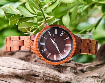May include: A wooden wristwatch with a brown face and Roman numeral hour markers. The watch has a wooden band and is set against a backdrop of green leaves and a piece of driftwood. The watch hands point to the time.