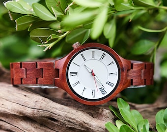 May include: A wooden wristwatch with a round face and a brown wooden band. The watch face is white with black Roman numerals and black hands, with a red second hand. The watch is displayed on a piece of wood with green leaves in the background.