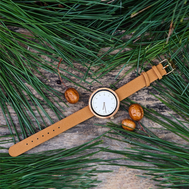 K&ouml;nnte beinhalten: Eine hellbraune Lederuhr mit hellem Holzgesicht und wei&szlig;em Zifferblatt. Die Uhr liegt auf einer Holzoberfl&auml;che mit gr&uuml;nen Kiefernnadeln und kleinen braunen Samenkapseln. Das Uhrenarmband ist hellbraun und hat eine goldene Schnalle.