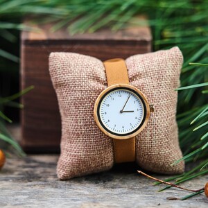 K&ouml;nnte beinhalten: Eine Holzarmbanduhr mit hellbraunem Lederarmband und wei&szlig;em Zifferblatt. Die Uhr wird auf einem kleinen, braunen Jutekissen pr&auml;sentiert. Die Uhr hat schwarze Zahlen und Zeiger. Der Hintergrund umfasst eine Holzkiste und gr&uuml;ne Tannennadeln.