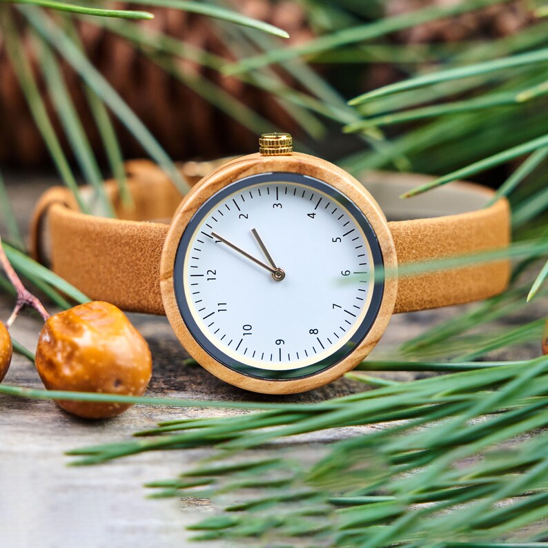 K&ouml;nnte beinhalten: Eine Holzarmbanduhr mit hellbraunem Lederarmband. Das Zifferblatt ist wei&szlig; mit schwarzen Ziffern und Zeigern. Die Uhr hat eine goldfarbene Krone und eine schwarze L&uuml;nette. Umgeben von gr&uuml;nen Kiefernnadeln und einem braunen Lederarmband.