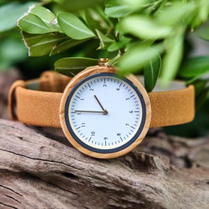 K&ouml;nnte beinhalten: Eine Holzarmbanduhr mit wei&szlig;em Zifferblatt, schwarzen und goldenen Akzenten und einem braunen Lederarmband. Die Uhr hat ein minimalistisches Design mit klarer Zeitanzeige. Die Uhr ist vor dem Hintergrund gr&uuml;ner Bl&auml;tter und Holz platziert.