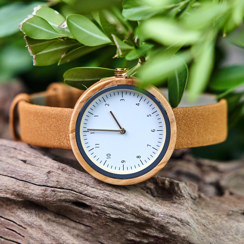 K&ouml;nnte beinhalten: Eine Holzarmbanduhr mit wei&szlig;em Zifferblatt, schwarzen und goldenen Akzenten und einem braunen Lederarmband. Die Uhr hat ein minimalistisches Design mit klarer Zeitanzeige. Die Uhr ist vor dem Hintergrund gr&uuml;ner Bl&auml;tter und Holz platziert.