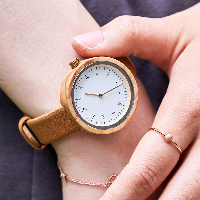 K&ouml;nnte beinhalten: Eine Holzarmbanduhr mit hellbraunem Lederarmband. Das Zifferblatt der Uhr ist wei&szlig; mit schwarzen Ziffern und goldfarbenen Zeigern. Die Uhr ist rund und hat eine goldfarbene Krone. Ein goldener Ring mit einem kleinen Stein befindet sich am Finger.