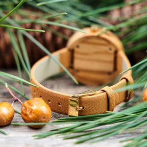 K&ouml;nnte beinhalten: Nahaufnahme einer Holzuhr mit einem hellbraunen Lederarmband und einer goldenen Schnalle. Das Zifferblatt der Uhr ist aus Holz. Die Uhr ist von Gr&uuml;nzeug und kleinen, runden, orangebraunen Beeren umgeben.