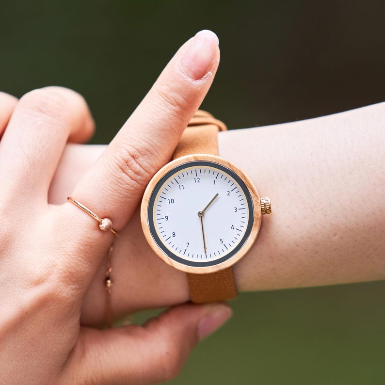 K&ouml;nnte beinhalten: Eine runde Holzuhr mit wei&szlig;em Zifferblatt und schwarzen Markierungen. Die Uhr hat ein braunes Lederarmband und goldfarbene Akzente. Das Zifferblatt zeigt die Zahlen 1 bis 12 an. Die Uhr wird am Handgelenk getragen.
