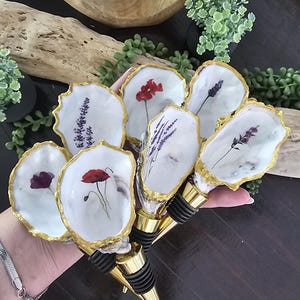 May include: A selection of decorative bottle stoppers. Each stopper features a white oyster shell design with a gold trim, displaying pressed flowers such as lavender and poppies. The stoppers have gold conical tips and black rubber seals.