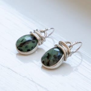 May include: A pair of drop earrings featuring faceted, oval-shaped green and black speckled stones. Each stone is encased in a silver-colored metal setting with decorative wire wrapping at the top, connected to a simple silver-colored ear wire hook.