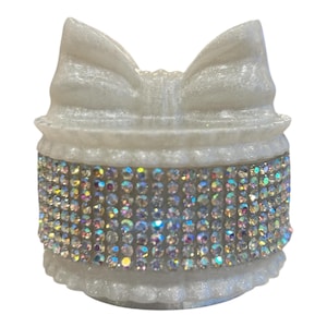 May include: A white, decorative trinket box with a bow-shaped lid. The box is adorned with iridescent rhinestones around the middle. The box has a scalloped base and a textured, pearlescent finish.