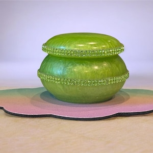May include: A lime green decorative jar with a lid, adorned with sparkling green rhinestones. The jar sits on a cloud-shaped mat with a gradient of pink, green, and yellow. The jar is a decorative item.