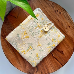 May include: A beige fabric pouch with a floral embroidery design in yellow and green. The pouch has a button closure and a fabric loop.