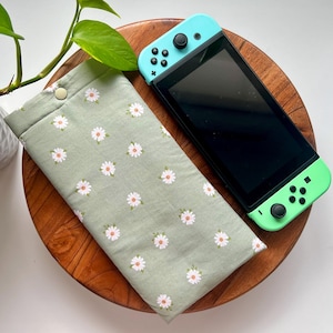 May include: A green and white floral fabric case for a Nintendo Switch console. The case has a button closure and a daisy print.