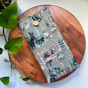 May include: A gray fabric case with a whimsical pattern featuring black cats in witch hats, cauldrons, crescent moons, and cupcakes. The case is secured with a wooden button and a loop closure. The case is on a wooden surface.