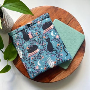 May include: A teal blue e-reader cover with a black cat and floral pattern. The cover features the text "The Charmed & Practical Potions".