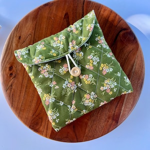 May include: A quilted green fabric pouch with a floral pattern in pink, yellow, and white. The pouch has a flap closure secured by a wooden button and a white cord loop. The pouch is on a round wooden surface.
