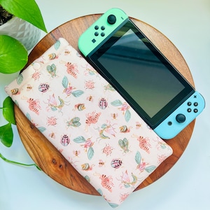 May include: A light pink fabric case with a repeating insect and floral pattern, next to a mint green and blue handheld gaming console. The case is rectangular and appears to be made of soft fabric. The console is black with mint green controllers.
