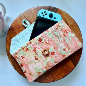 May include: A light blue Nintendo Switch console rests inside a pink floral patterned case with a wooden button closure. The case is open, revealing a light blue interior. The case sits on a round wooden tray.