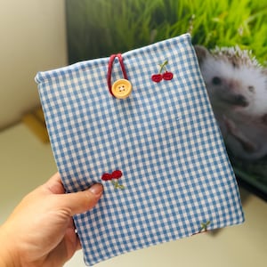 May include: A blue and white gingham pattern fabric case with embroidered red cherries and a wooden button closure. The case has a red cord loop and is designed to hold a tablet or e-reader.