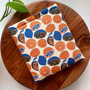 May include: A white fabric book cover with a repeating pattern of orange and blue citrus fruit slices. The cover has a single orange button.