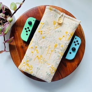 May include: A cream-colored embroidered case with a button closure, featuring yellow and green floral designs, cradles a mint green and a light blue game controller. The case rests on a wooden tray, with a potted plant in the background.
