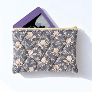 May include: A quilted fabric pouch in a dark gray color featuring a repeating light beige floral pattern. The pouch has a gold-toned zipper closure at the top and contains a purple e-reader with the word Kindle visible on the screen.