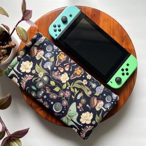 May include: A teal and black Nintendo Switch console rests on a wooden tray next to a fabric case. The case features a dark blue background with a floral and insect pattern, including butterflies, mushrooms, and flowers. A small potted plant is visible.