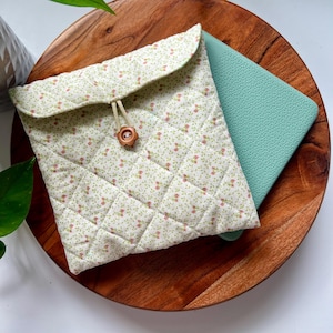 May include: A quilted fabric case with a floral pattern in green and pink, secured with a wooden button closure. The case is on a wooden tray, with a teal item partially visible inside.