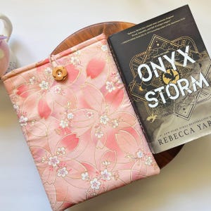 May include: A pink floral book sleeve with a wooden button closure next to a book titled "ONYX STORM" by Rebecca Yarros. The book sleeve features a pattern of pink cherry blossoms with gold accents.