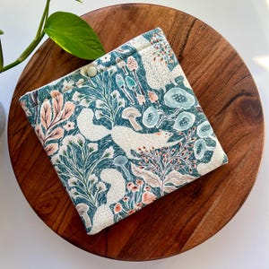May include: A white fabric pouch with a teal and white floral pattern featuring foxes and mushrooms. The pouch has a white button closure.