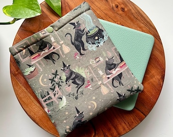 May include: A green fabric pot holder with a black cat and witch hat design. The pot holder has a green button and is on a wooden surface.