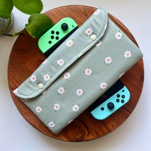 May include: A green and white floral fabric case for a Nintendo Switch console. The case has a snap closure and is shown with two teal and green controllers.