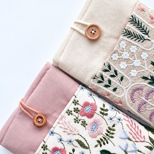 May include: Two fabric pouches with button closures. One pouch is cream-colored with green and white floral embroidery. The other is dusty pink with a white panel featuring pink, blue, and green floral embroidery. Both have matching cord loops.