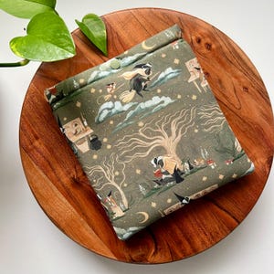 May include: A square fabric pouch in olive green featuring a repeating pattern of badgers, crescent moons, stars, and trees. The pouch has a fold-over top secured with a single light green snap button and displays illustrations of badgers reading books.