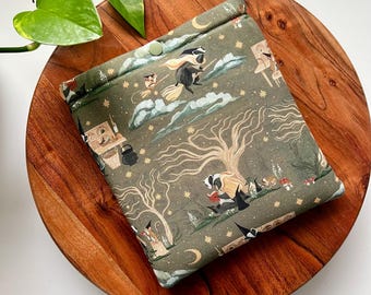 May include: A square fabric pouch in olive green featuring a repeating pattern of badgers, crescent moons, stars, and trees. The pouch has a fold-over top secured with a single light green snap button and displays illustrations of badgers reading books.