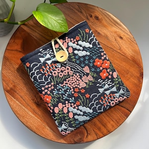 Könnte beinhalten: Eine dunkelblaue Stofftasche mit einem floralen Stickmuster in Rosa-, Orange- und Weißtönen. Die Tasche hat einen Verschluss aus Holz und einen weißen Kordelzug.