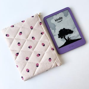 May include: A quilted fabric pouch in a light pink hue featuring a repeating pattern of small dark pink strawberries, paired with a purple e-reader displaying the word kindle and a graphic of a person sitting under a tree.