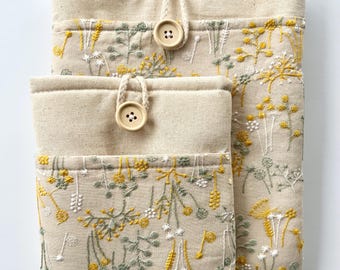 May include: Two beige linen pouches with a floral embroidery design in shades of yellow and green. The pouches have a drawstring closure and a button detail.
