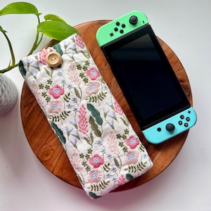 Può includere: Una custodia in tessuto bianco con un ricamo floreale e una chiusura a bottone. La custodia è appoggiata su una superficie di legno accanto a una console Nintendo Switch color turchese.