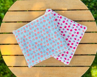 Pode incluir: Duas bolsas com zíper com um padrão de morangos. Uma é azul clara com morangos vermelhos e rosa, a outra branca com morangos rosa. As bolsas estão em uma mesa redonda de madeira.