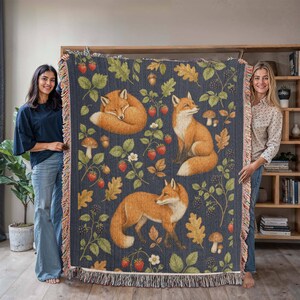 Könnte beinhalten: Eine gewebte Wohndecke mit dunkelblauem Hintergrund und einem wiederkehrenden Muster aus orangefarbenen Füchsen, roten Erdbeeren, Brombeeren, braunen Eicheln, Pilzen und grünen Eichenblättern. Die Decke hat an allen Seiten einen Fransenrand.