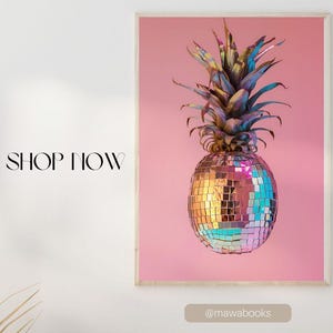 Könnte beinhalten: Ein gerahmtes Poster mit einer Discokugel-Ananas vor rosa Hintergrund. Die Ananas hat eine verspiegelte Oberfläche und blau-grüne Blätter. Das Bild wird an einer weißen Wand über einer Holzkonsolentisch präsentiert.