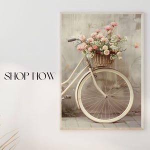 Puede incluir: Un póster enmarcado con una bicicleta vintage con una cesta de flores rosas y blancas. El póster se muestra encima de una consola de madera con cestas decorativas y una lámpara. El texto en la pared dice "Decora tus paredes", "Compra ahora" y "Pósters".