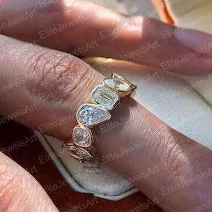 May include: A gold ring with a sequence of clear, differently shaped gemstones. The gemstones include a pear shape, emerald cut, and other geometric forms. The ring is set in a gold band, creating a modern and elegant design.